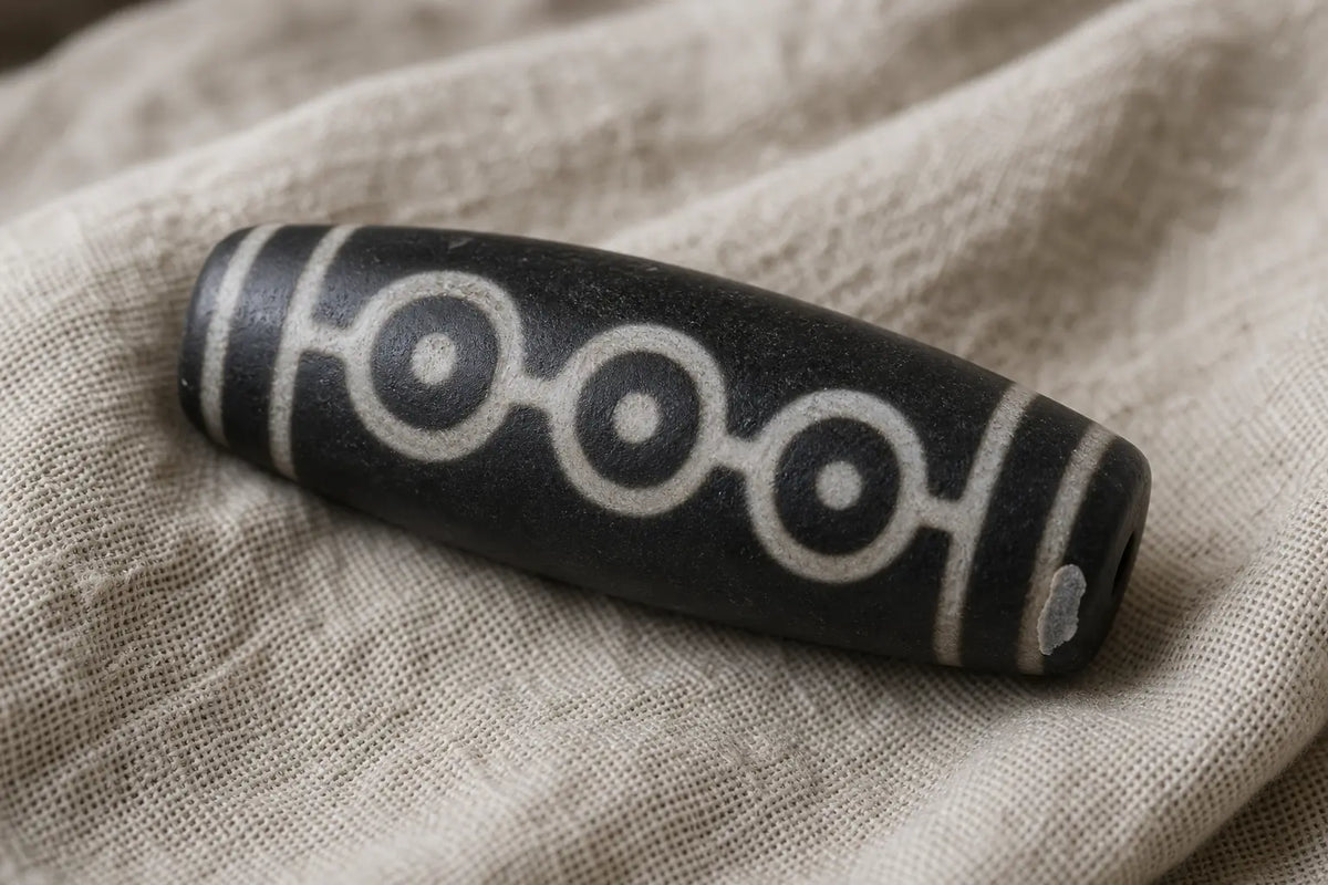 A barrel-shaped black dzi bead with three white eyes linked by white bands, resting on coarse undyed linen, with a small chip at one end showing the pale stone beneath