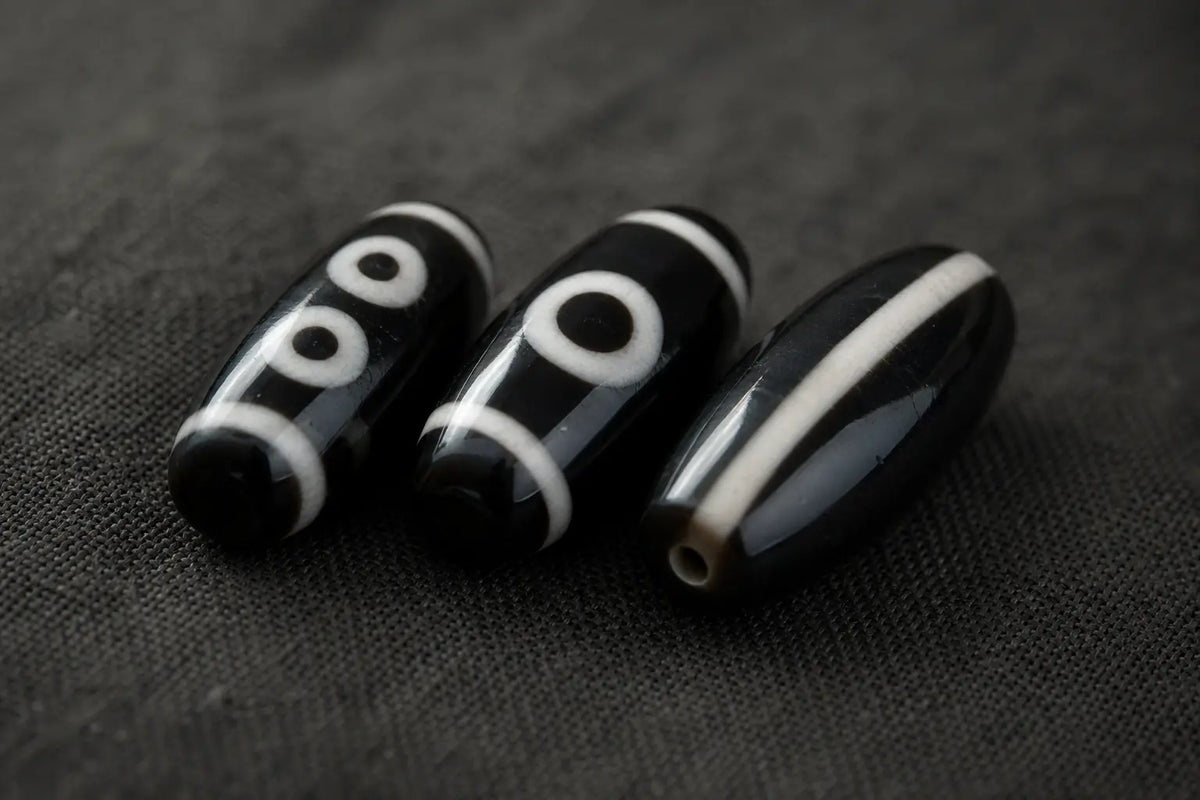 Three black-and-white dzi beads resting on dark linen, each with white eye circles and line work on a dark ground, soft daylight raking across the stone