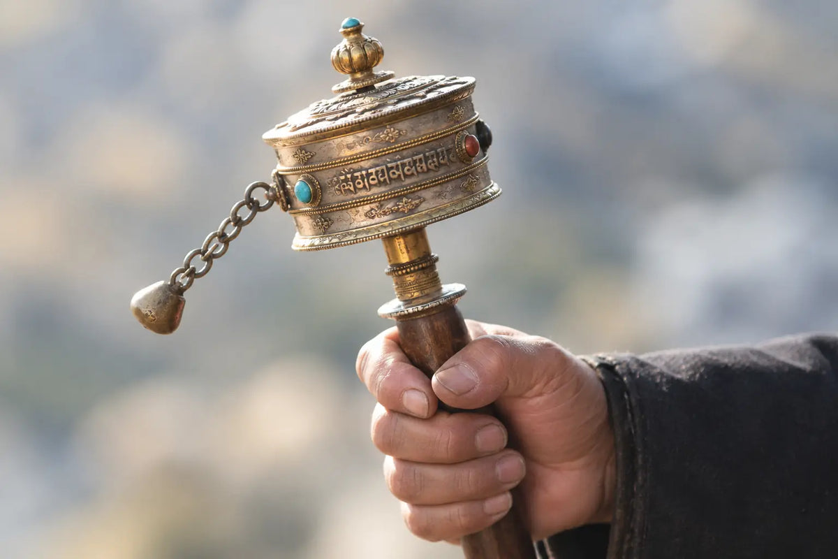 A hand holding the wooden grip of a hand-held Tibetan prayer wheel, its silvered and gilt drum set with turquoise and coral around an engraved mantra band, a brass counterweight hanging from a chain at its side