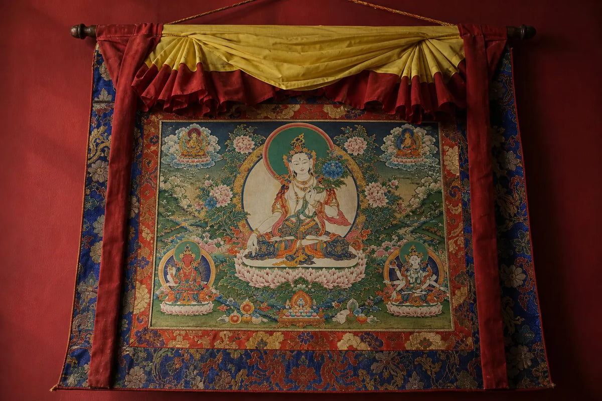 A finished Tibetan thangka hung from a dark rod, a serene green-haloed deity painted on cotton inside a deep-red and yellow silk brocade border and pleated canopy