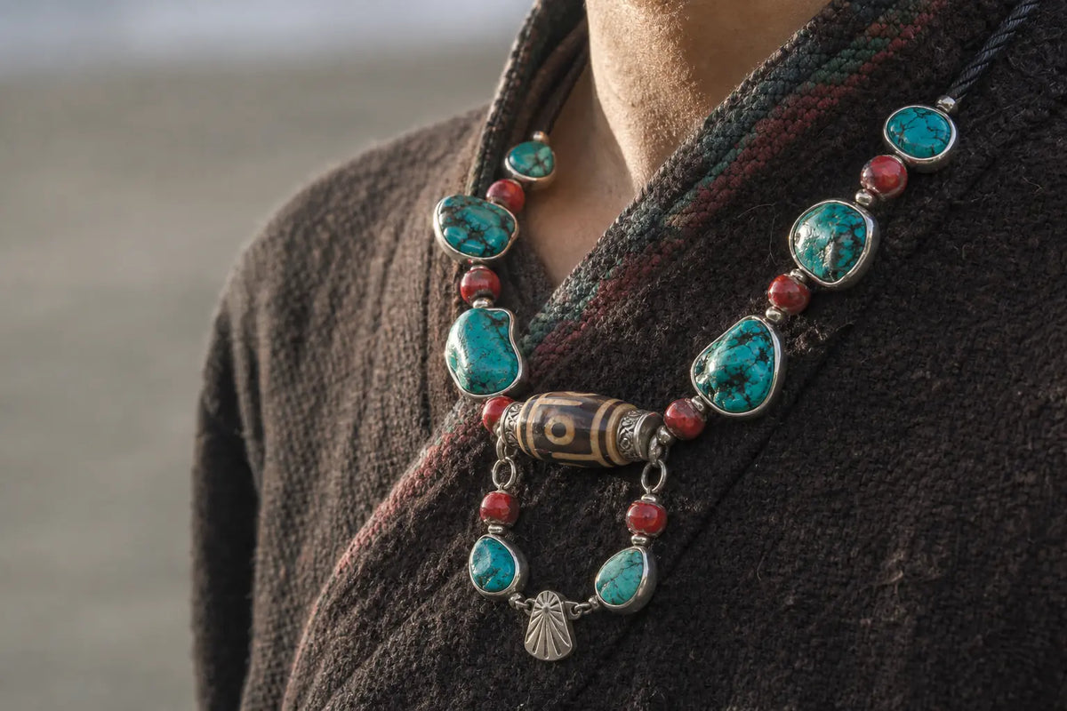 A Tibetan silver necklace with rough turquoise cabochons, red coral beads and a banded agate dzi bead, worn over a dark wool robe, cropped at the chest with no face in frame